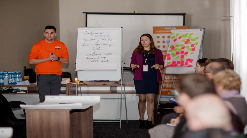 A man and a woman stand near flipcharts and present their work to the audience